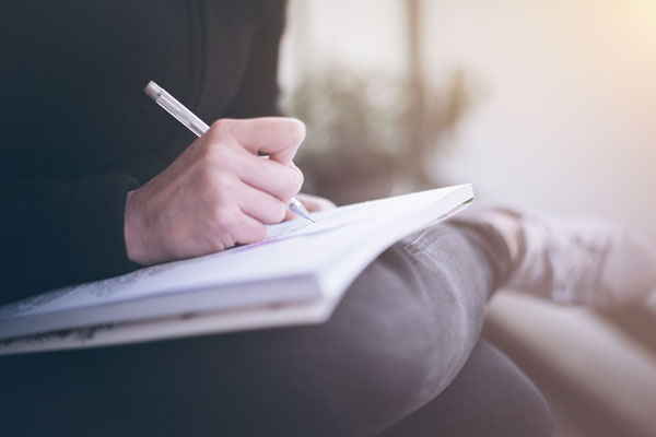 An image showing a hand writing in a notebook which is rested upon their knee